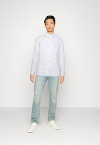 Ανοιχτό μπλε μακρυμάνικο πουκάμισο με κουμπιά και μικρό λογότυπο, συνδυασμένο με ανοιχτόχρωμο τζιν και λευκά sneakers, σε απλό φόντο.