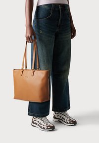 Bruine leren tote bag met gouden hardware, rechthoekige vorm en dubbele handvatten; gedragen met oversized blauwe spijkerbroeken en multikleuren sneakers.