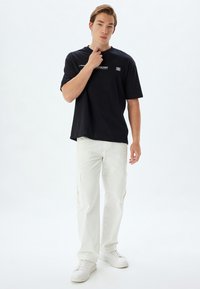Camiseta negra de manga corta con un diseño gráfico sutil, combinada con pantalones cargo blancos de corte holgado que cuentan con bolsillos laterales y cremalleras.