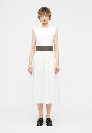 Žena stojaca čelom dopredu, oblečená v bielych midi šatách bez rukávov s tmavým pásom v páse a čiernymi mokasínami, na jednoduchom pozadí.