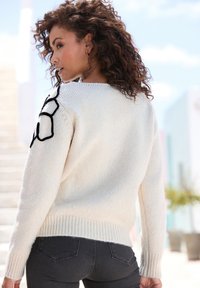 Cremfarbener Pullover mit geripptem Saum, der ein schwarzes Blumenmuster auf der Schulter aufweist, kombiniert mit dunkelgrauen, eng anliegenden Jeans.