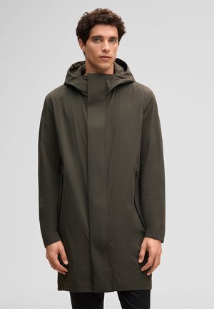 Młody mężczyzna w oliwkowozielonym, do kolan, przeciwdeszczowym płaszczu z kapturem, z ukrytym przednim zamkiem błyskawicznym i bocznymi kieszeniami, stojący na jednolitym tle.