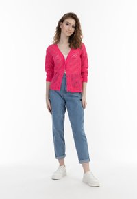Rosa stickad cardigan med diamantmönster, knäppning med knappar, ribbade muddar; kombinerad med högmidjade ljusblå jeans och vita sneakers.