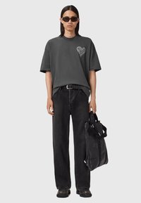 Camiseta gris de gran tamaño con gráfico de corazón, pantalones de mezclilla negros y zapatos negros gruesos. La modelo lleva una bolsa tote negra con detalle de logo.