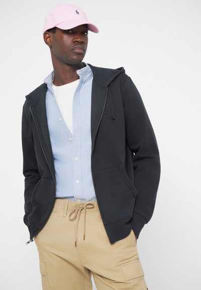 Μαύρη κουκούλα με φερμουάρ και τσέπες, φορεμένη πάνω από ένα ανοιχτό μπλε πουκάμισο με κουμπιά και κρεμ cargo παντελόνι. Ροζ καπέλο φορεμένο στο κεφάλι.