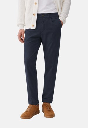 Uomo che indossa chinos blu navy scuro con risvolti, camicia bianca, cardigan crema con bottoni in legno e sneakers in pelle scamosciata marrone.