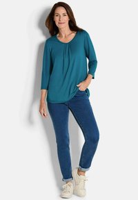 Tee-farbene Langarmbluse mit Rundhalsausschnitt und Falten, kombiniert mit blauen Jeans und weißen Sneakern. Einfacher, legerer Schnitt und leichtes Material.