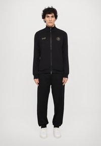 Svart zip-jacka med hög krage, guld EA7-logga och matchande svarta joggers; tyget ser slätt ut; bärs med vita sneakers.