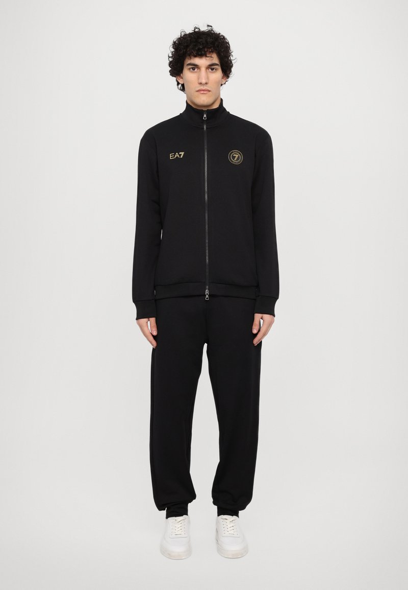 Svart zip-jacka med hög krage, guld EA7-logga och matchande svarta joggers; tyget ser slätt ut; bärs med vita sneakers.