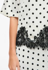 Tissu blanc à pois noirs, orné d'une bordure en dentelle noire en bas. La dentelle présente des motifs floraux et une finition texturée.