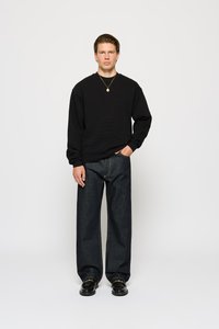Sweatshirt noir texturé à manches longues, associé à un jean en denim foncé ample. Le modèle se tient droit, les mains dans les poches, portant des chaussures noires.