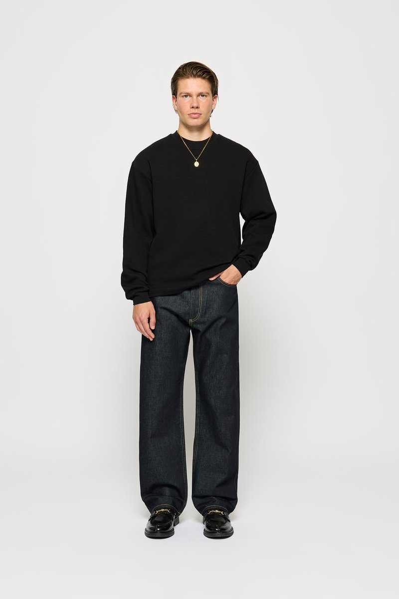 Sweatshirt noir texturé à manches longues, associé à un jean en denim foncé ample. Le modèle se tient droit, les mains dans les poches, portant des chaussures noires.