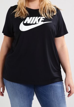 Personne blonde portant un T-shirt noir Nike et un jean bleu, debout devant un fond clair uni.
