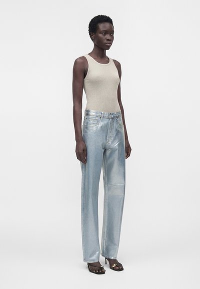 Γυναίκα με μπεζ αμάνικο τοπ, μεταλλικό ανοιχτό μπλε τζιν με ίσια πόδια και μαύρες ανοιχτές γόβες με χρυσή λεπτομέρεια, στέκεται μπροστά σε λευκό φόντο.