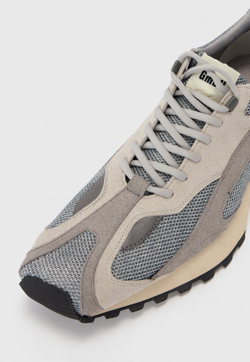Chaussure de sport beige et grise avec une tige en mesh, des accents en daim et une semelle en caoutchouc. Présente des motifs texturés et un col rembourré.
