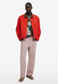 Rote Canvasjacke mit braunem Kragen, darunter ein blumengemustertes Hemd, hellrosa Hosen und braune Slipper.