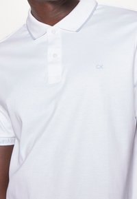 Vit pikétröja med mjuk textur, tvåknappslåsning och ljusblå "CK"-logotyp på bröstet. Har en diskret krage.