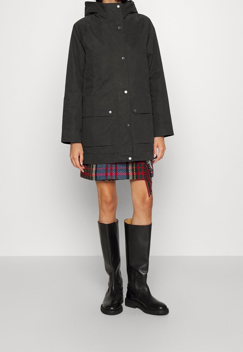 Black waterproof parka with button closure, two front pockets, and adjustable hood, paired with a plaid skirt and knee-high black boots.