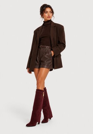 Frau trägt einen dunkelbraunen Blazer, Rollkragenpullover, Leder-Shorts und hohe burgunderrote Wildlederstiefel und steht vor einem schlichten hellen Hintergrund.