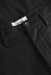 Schwarze Hose mit glattem Stoff, ausgestattet mit einem Knopfverschluss vorne, Gürtelschlaufen und einem GABBA-Label.