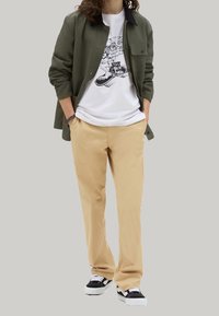 Camicia verde oliva con bottoni, t-shirt bianca con stampa grafica, pantaloni beige, sneakers nero e bianco. Il tessuto sembra liscio; il design presenta una stampa grafica.