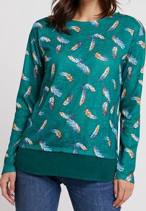 Turkooise top met lange mouwen met een meerkleurig veerpatroon, gedragen over een turkooise gelaagde zoom, gecombineerd met blauwe denim jeans.