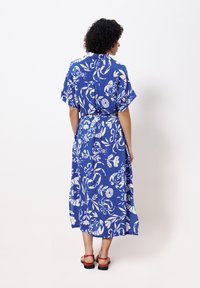 Maxikleid in Blau mit weißen Blumenmustern, kurzen Ärmeln, Gürtelakzent in der Taille und lockerer Passform. Getragen mit roten Sandalen.