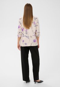 Blazer beige à motif floral avec les manches retroussées, associé à un pantalon noir et des talons noirs. Présente des accents floraux violets et bleus.