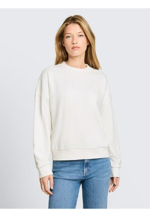 Junge Frau mit blondem Haar, die einen schlichten weißen Langarmpullover und blaue Jeans trägt, steht vor einem hellgrauen Hintergrund.