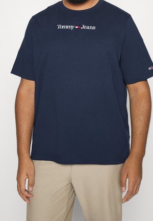 Mężczyzna w granatowej koszulce Tommy Jeans z logo na piersi i lewym rękawie, w połączeniu z beżowymi spodniami na jednolitym tle.