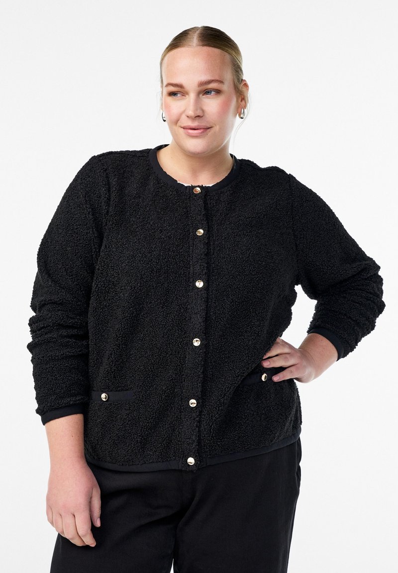 Sieviete melnā teksturētā jaka ar zeltītām pogām, roka uz gurniem, un melnās bikses uz vienkārša baltā fona.