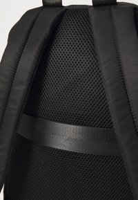 Černý batoh s texturovanou síťovinou, hladkým materiálem a horizontálním popruhem z pleteného materiálu ve středu pro větší oporu.