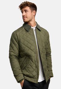 Quilted olivengrøn jakke med krave, med diagonale mønstre, lynlås lukning og sidelommer, sat mod en ensfarvet baggrund.