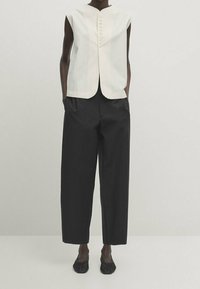 Biała bezrękawnik z okrągłym dekoltem i guzikami, w zestawieniu z czarnymi spodniami z szerokimi nogawkami oraz czarnymi plecionymi butami wsuwanymi.