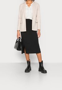 Blazer beige, chemise blanche à pois noirs, jupe noire jusqu'au genou avec une fente, bottines noires, portant un sac à main noir texturé.