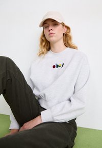 Sudadera gris con el logo "obey" en multicolor, combinada con pantalones de pana verde oliva. Modelo lleva una gorra beige en interiores.