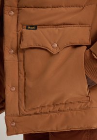 Casaco isolante castanho com um bolso horizontal, botões de pressão e textura macia. Etiqueta da marca visível no bolso.