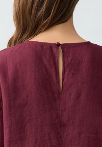 Top in lino borgogna con apertura a chiave sul retro e chiusura a bottone, caratterizzato da una consistenza morbida e un design semplice e svasato.
