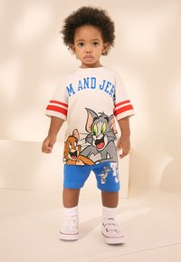 Vitt kortärmat t-shirt med röda ränder och Tom och Jerry-grafik; kombineras med blå shorts med en tecknad logotyp; vita sneakers.