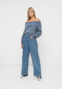 Μπλε πλεκτό πουλόβερ με ανοιχτούς ώμους και ριμπ λαιμό, συνδυασμένο με ψηλόμεσα, φαρδιά τζιν και μαύρα πάνινα αθλητικά παπούτσια.