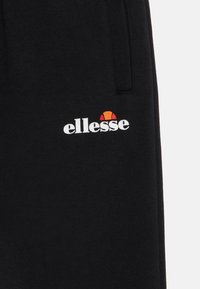 Crna tkanina s bijelim "ellesse" logotipom i narančasto-crvenim polukrugom blizu šavova džepova odjevnog predmeta.