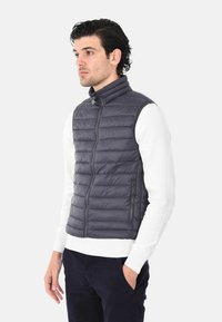 Gilet grigio imbottito con design a righe verticali, colletto alto, zip completa e tasche laterali, indossato sopra una camicia bianca a maniche lunghe.