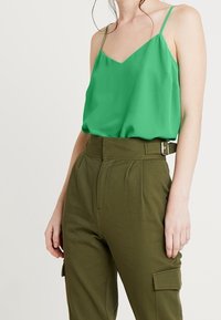 Blusa in seta verde, senza maniche, con scollo a V e spalline sottili, abbinata a pantaloni cargo verdi oliva a vita alta con cintura e tasche.