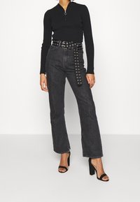 Haut noir à manches longues côtelé avec col et fermeture éclair, associé à un jean large taille haute gris foncé et des sandales noires à talons blocs.