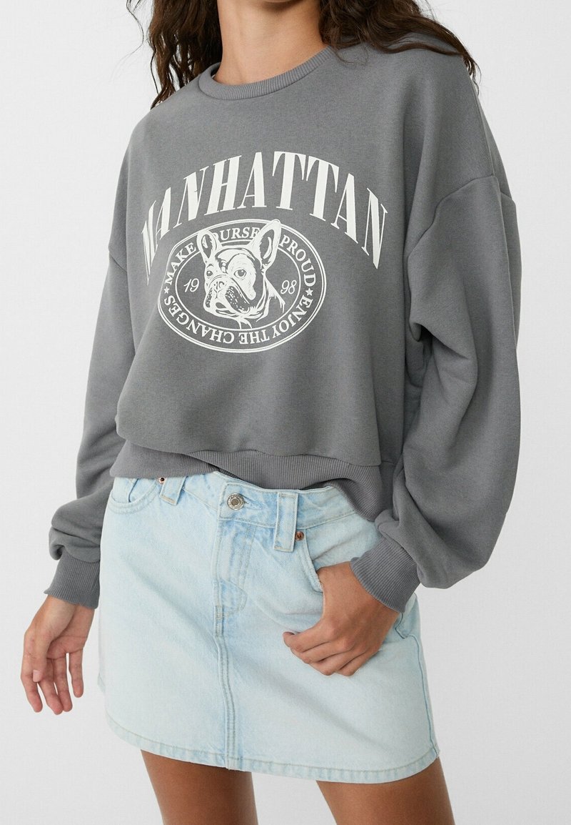 Grauer, kürzer geschnittener Sweatshirt mit einem weißen Grafikdruck eines Französischen Bulldoggen und Text, kombiniert mit einem hellblauen Jeansrock mit vorderem Knopfverschluss.