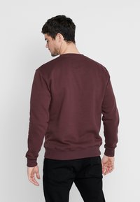 Mand iført en mørkebrun langærmet sweatshirt og sorte bukser, stående med ryggen til kameraet mod en hvid baggrund.