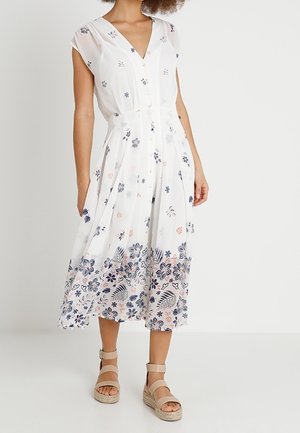 Kobieta w białej sukience midi bez rękawów z niebiesko-różowym kwiatowym wzorem i beżowych sandałach na platformie, stojąca na jednolitym tle.