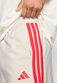 Αθλητικά σορτς σε ανοιχτό μπεζ χρώμα με τρεις κάθετες κόκκινες ρίγες στα πλαϊνά και μικρό κόκκινο λογότυπο στο κάτω αριστερό μ�έρος. Ορατή ελαστική ζώνη μέσης.