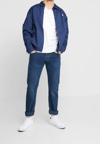 Námořnicky modrá větrovka s límcem, elastickými manžetami a bílým logem, doplněná bílým tričkem a ohrnutými tmavě modrými džínami, bílé tenisky.