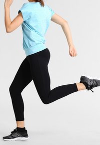 Top deportivo azul claro con mangas cortas, combinado con leggings negros. Los zapatos presentan un patrón de zigzag en blanco y negro. El sujeto está en movimiento.